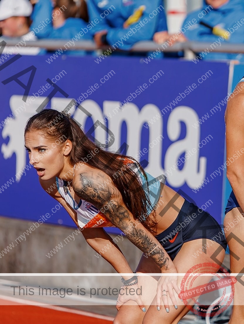 karolina-manasova-czechia-karolina_manasova-388 Karolína Maňasová, with long brown hair and an arm tattoo, bends forward catching her breath at a race; blue-jacketed people behind.