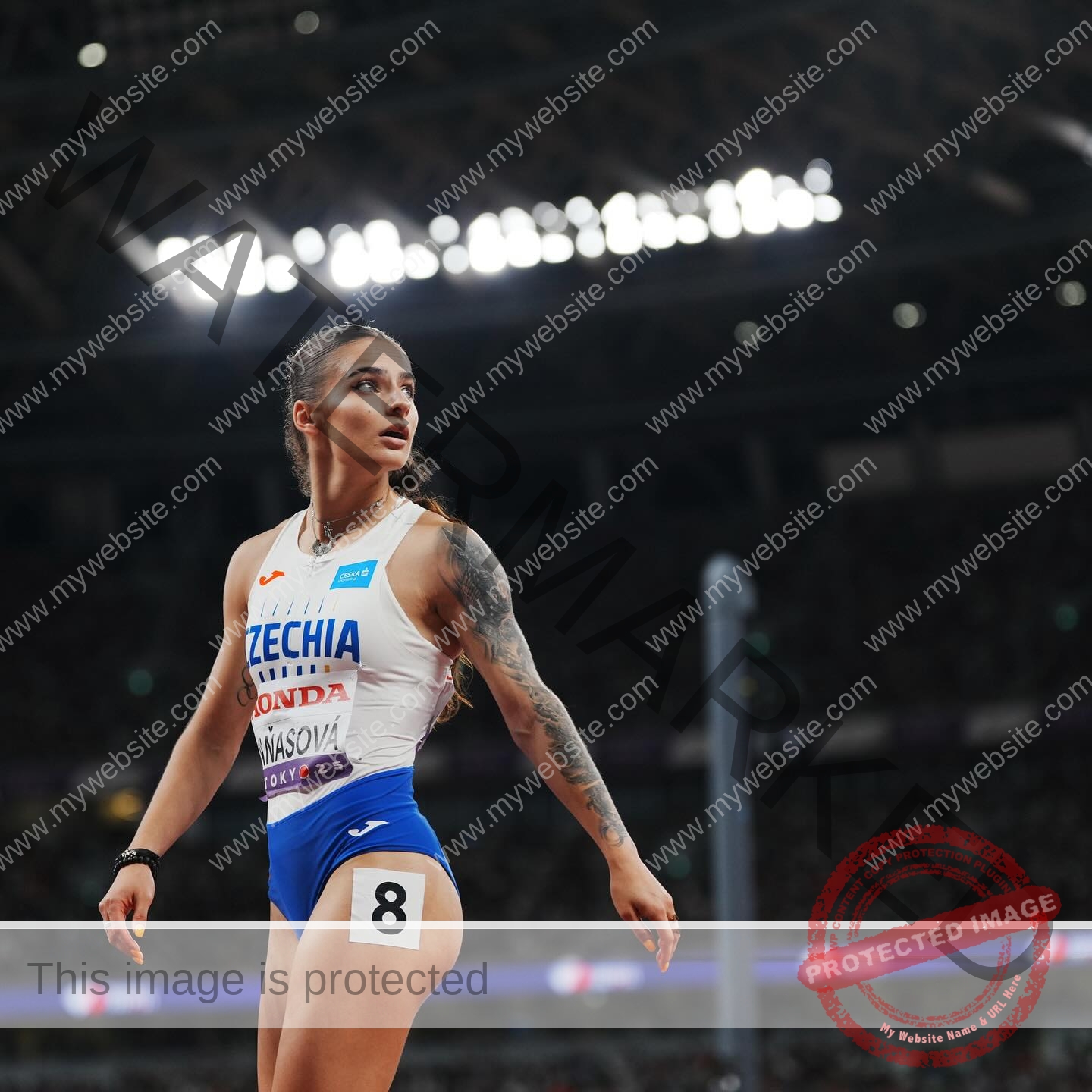 karolina-manasova-czechia-karolina_manasova-393 Karolína Maňasová, in a blue and white Czechia uniform with number 8 shorts, stands focused on a track; stadium lights shine behind.