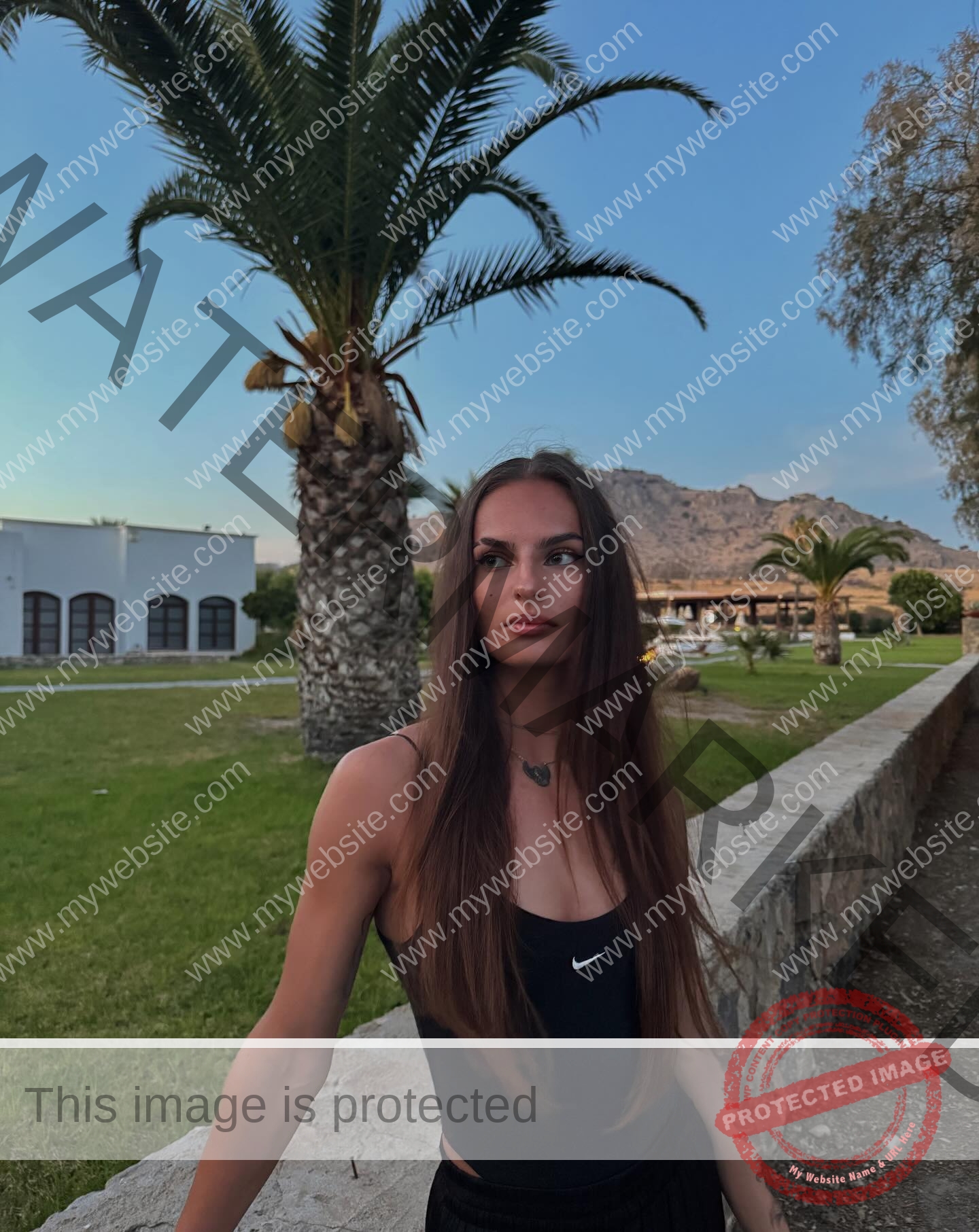 karolina-manasova-czechia-karolina_manasova-395 Karolína Maňasová with long brown hair in a black Nike tank top stands by a stone wall and palm trees at sunset, mountains behind.
