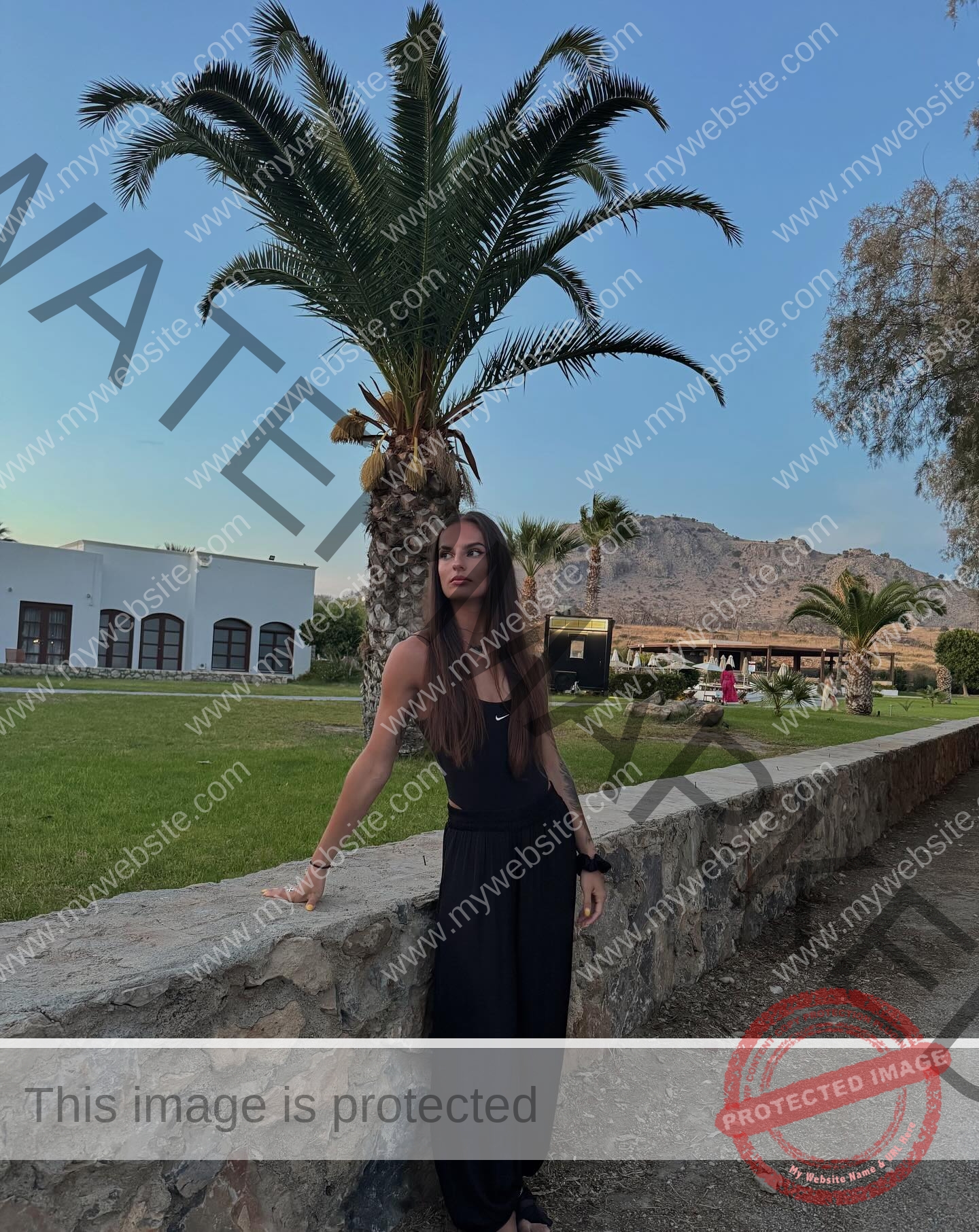 karolina-manasova-czechia-karolina_manasova-400 Karolína Maňasová in a black outfit stands by a stone wall, with palm trees, mountains, and arched-window buildings behind her.