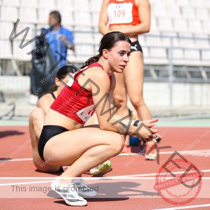 Paola Shyle, in a red top and black shorts, crouches on a running track, focused before a race. Others and seats are behind.