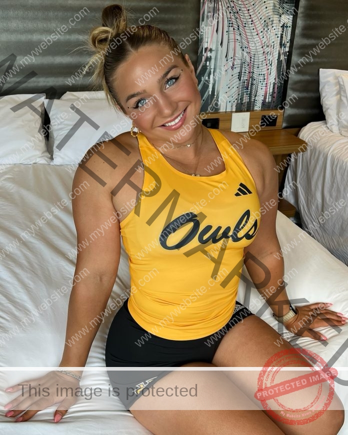 Kali Terza A woman with blonde hair in buns, wearing a yellow Kennesaw State Owls tank top and black shorts, smiles on a bed in a modern room.