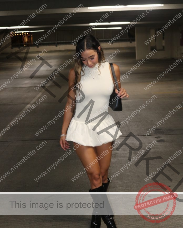 Katia Gallegos A woman in a white sleeveless dress and black knee-high boots walks alone in a dim parking garage, holding a black purse.