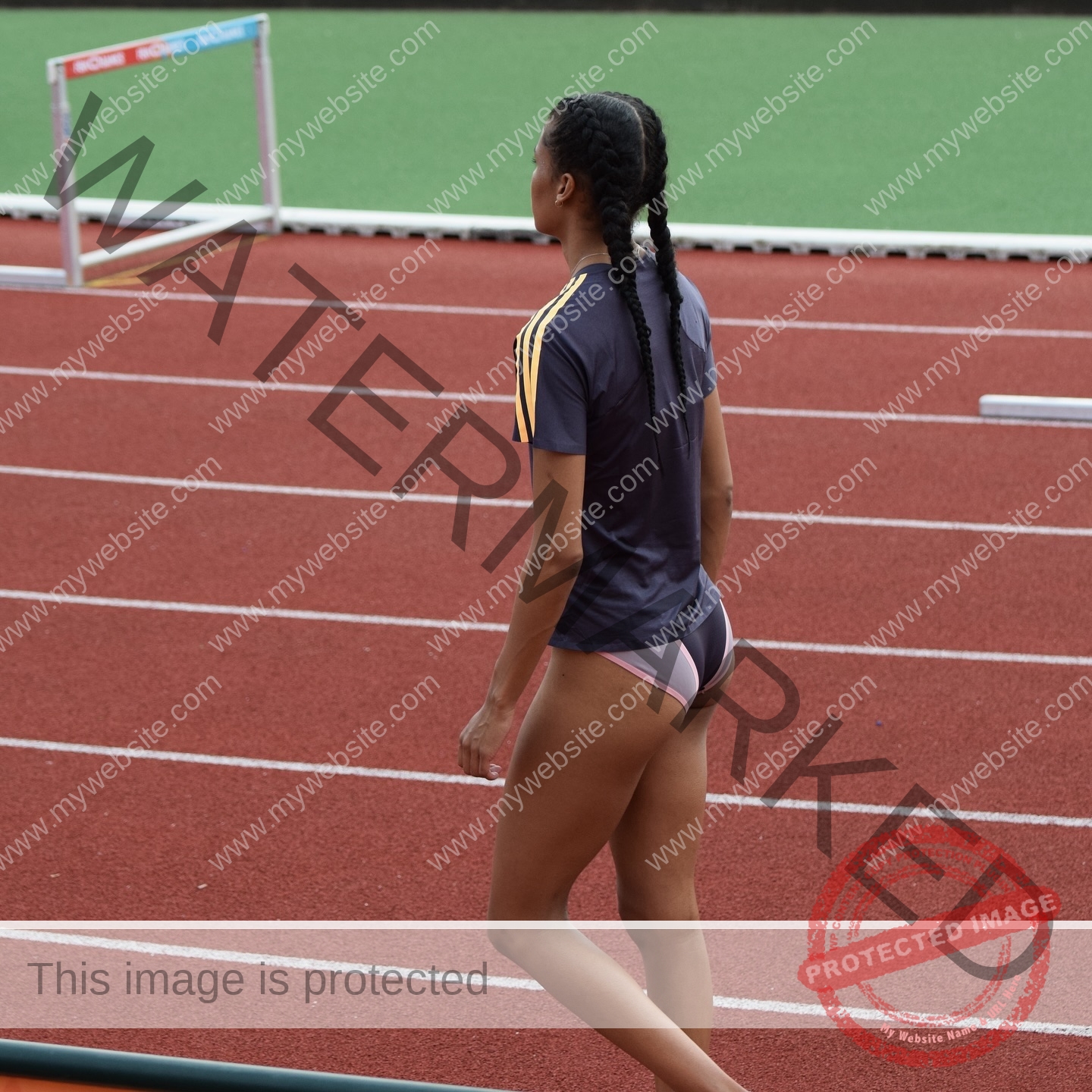abigail-noruwa-netherlands-abigail_noruwa-u20-x-00034 Abigail Noruwa, track star from the Netherlands, walks on a red track near a hurdle in dark gear with yellow stripes and sport briefs.