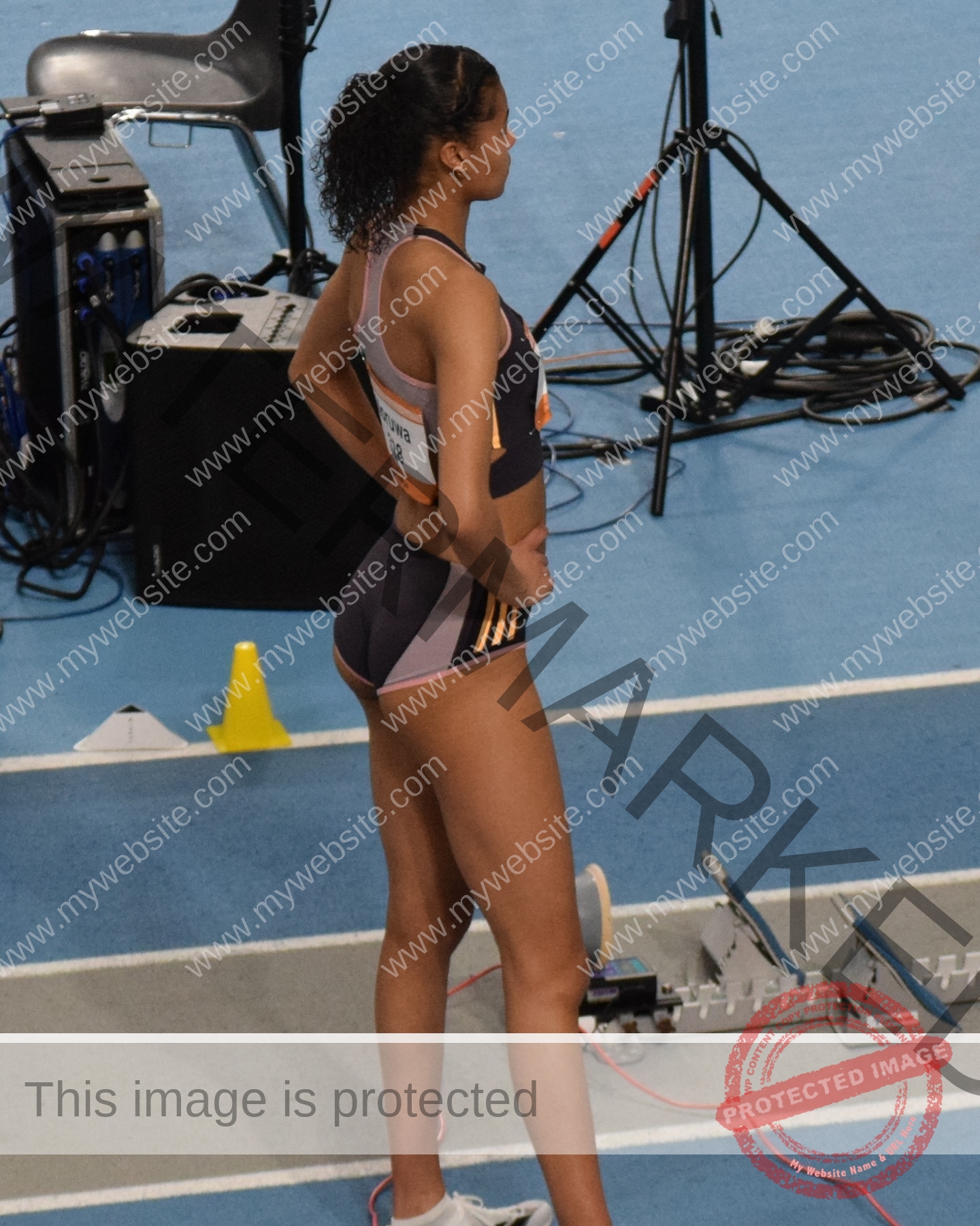 abigail-noruwa-netherlands-abigail_noruwa-u20-x-00057 Abigail Noruwa, track star from the Netherlands, stands hands on hips by starting blocks on an indoor track with gear behind her.