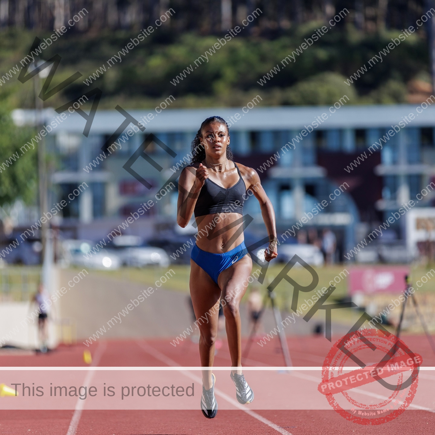 abigail-noruwa-netherlands-abigail_noruwa-u20-x-00061 Abigail Noruwa, track star from the Netherlands, sprints on an outdoor track in a black sports bra and blue shorts; blurred buildings and trees behind.