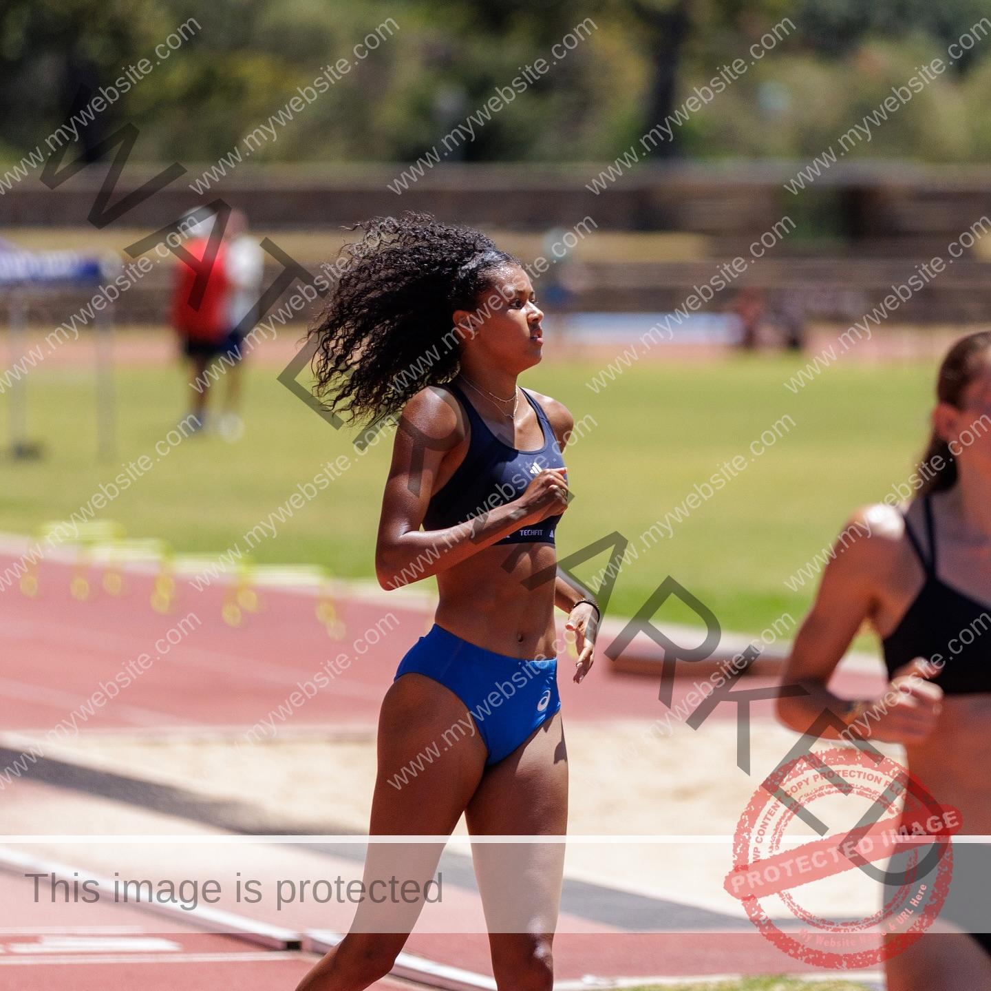 abigail-noruwa-netherlands-abigail_noruwa-u20-x-00070 Abigail Noruwa, track star from the Netherlands, runs in blue sportswear on a sunny track with other runners and hurdles behind.