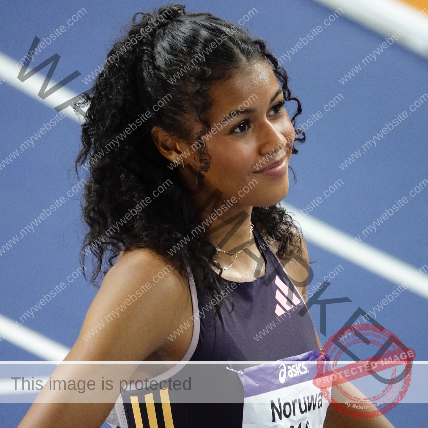 abigail-noruwa-netherlands-abigail_noruwa-u20-x-00072 Abigail Noruwa, track star from the Netherlands, stands on a blue indoor track in a dark top, smiling slightly and looking sideways.