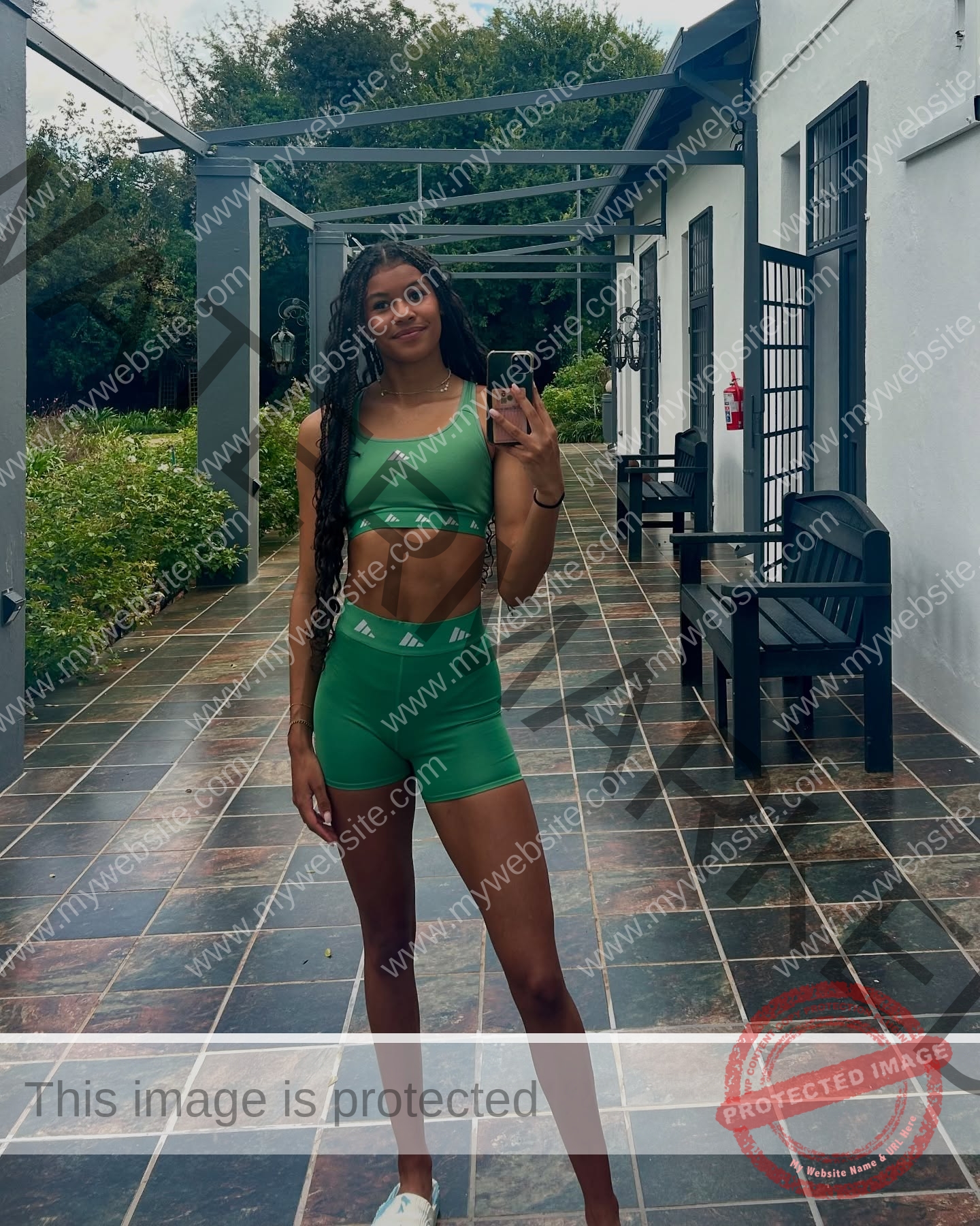 abigail-noruwa-netherlands-abigail_noruwa-u20-x-00082 Abigail Noruwa, track star from the Netherlands, wears a green sports set, smiling for a mirror selfie on a patio with plants behind.