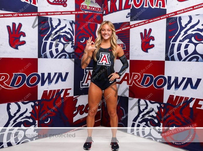 Addysan Prassas, a female athlete in a black Arizona Wildcats uniform, poses before a red, white, and blue Bear Down Nike backdrop, smiling and giving a hand sign.
