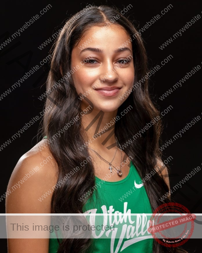 Adrian Powell A young woman with long wavy brown hair smiles softly, wearing a green Utah Valley track tank and cross necklace.
