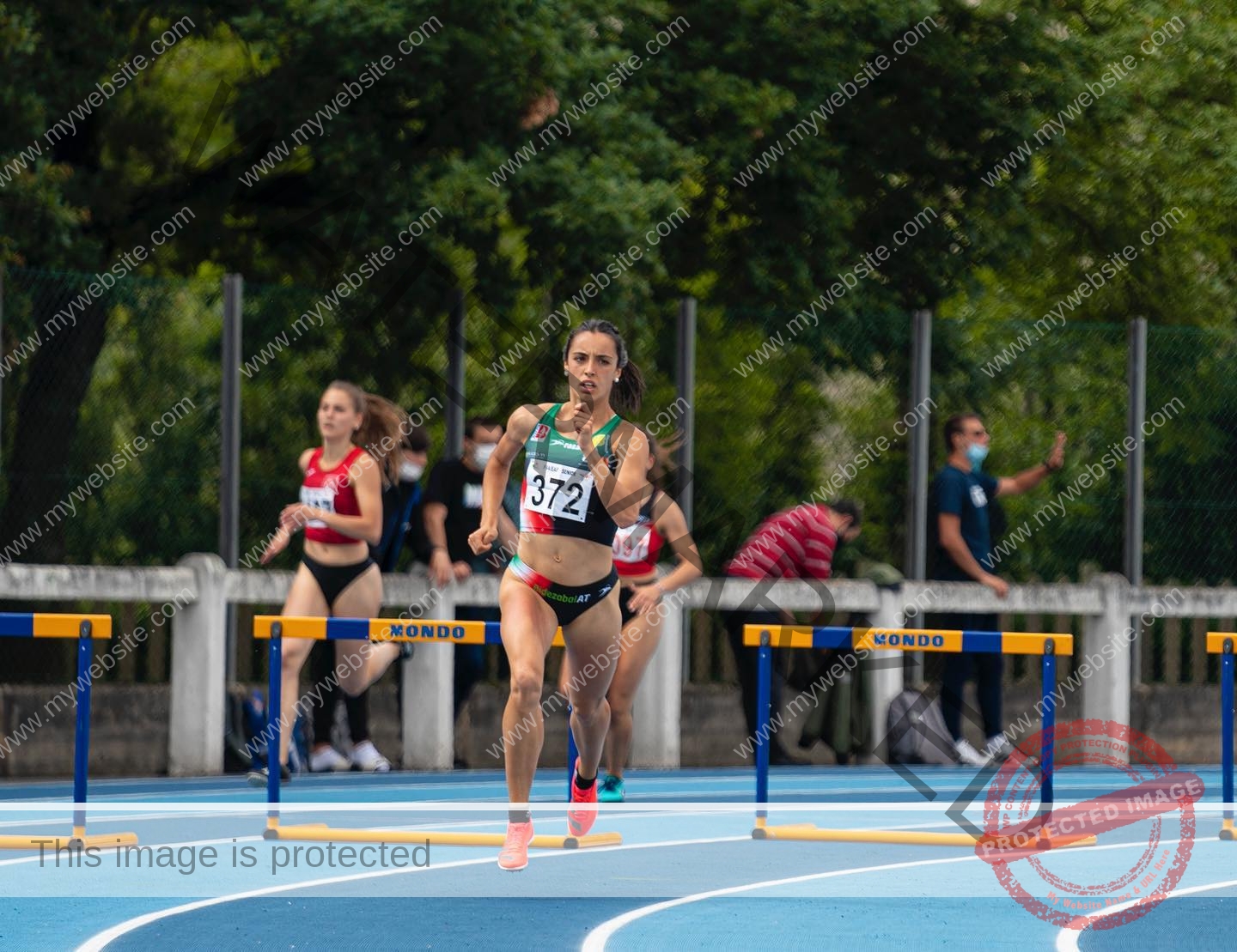 ainhoa-idoiaga-spain-ainhoidoiaga-00287 Ainhoa Idoiaga, track athlete from Spain, wearing bib 372 sprints ahead on a blue track while jumping hurdles; spectators behind.