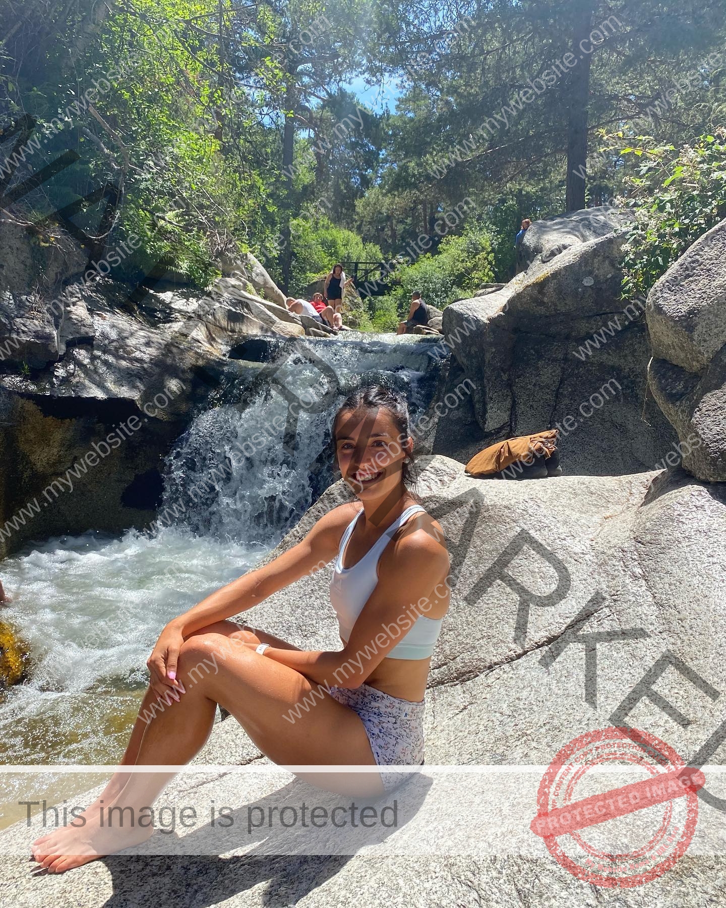 ainhoa-idoiaga-spain-ainhoidoiaga-00288 Ainhoa Idoiaga, track athlete from Spain, sits in a white bikini on a sunlit rock by a small waterfall in a forest; people relax nearby.