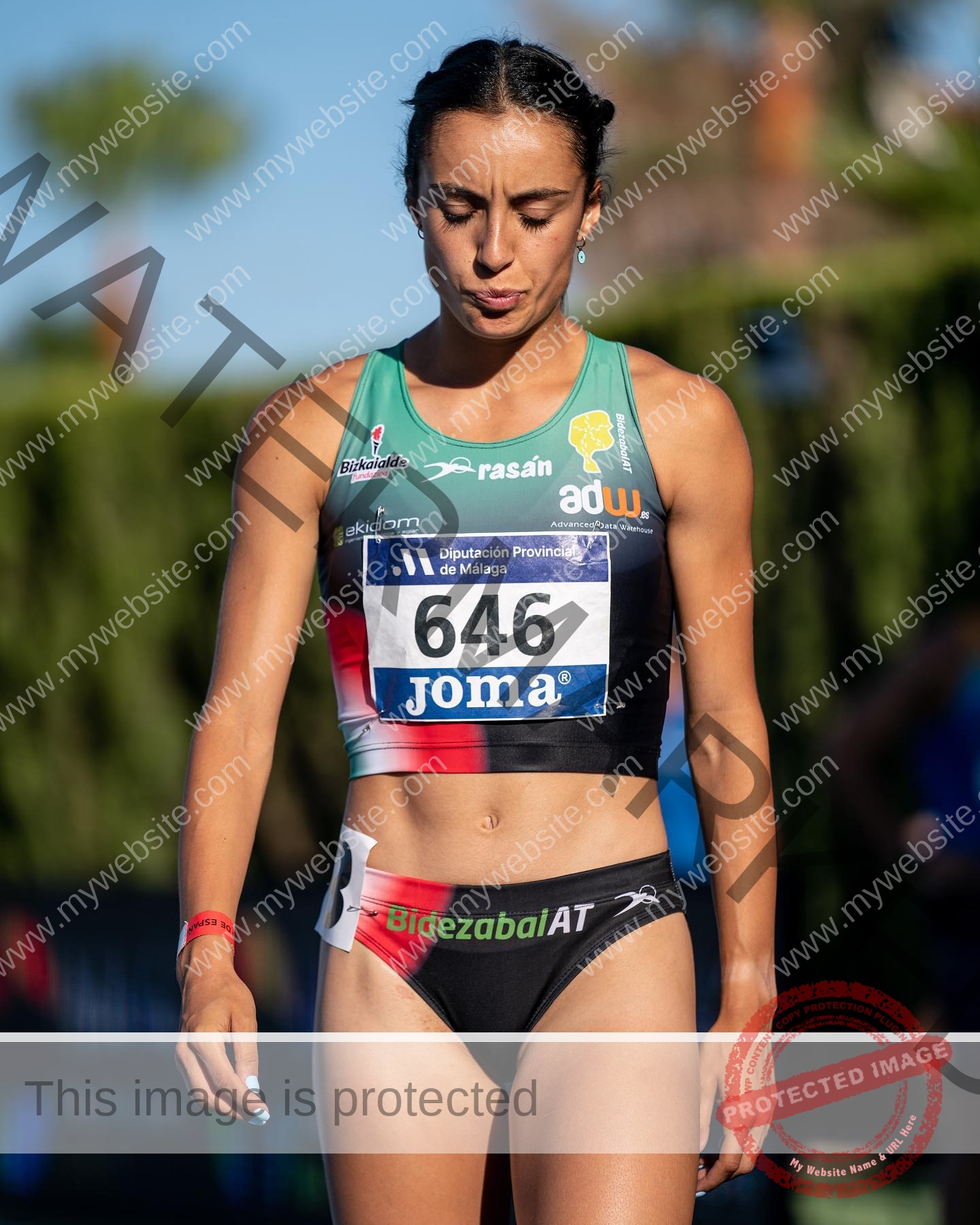 ainhoa-idoiaga-spain-ainhoidoiaga-00313 Ainhoa Idoiaga, track athlete from Spain, stands outdoors in sunlight wearing bib 646, focused, with trees and people behind.