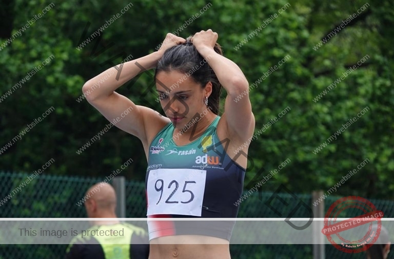 ainhoa-idoiaga-spain-ainhoidoiaga-00330 Ainhoa Idoiaga, track athlete from Spain, wearing race bib 925 ties her hair near a fence with greenery; athletes blurred behind.