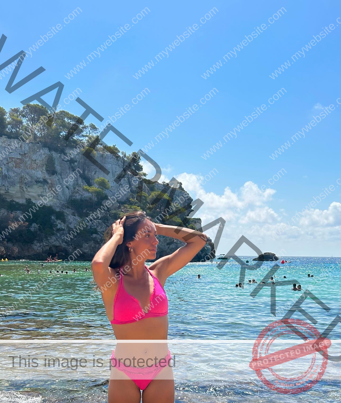 ainhoa-idoiaga-spain-ainhoidoiaga-00335 Ainhoa Idoiaga, track athlete from Spain, stands in a pink bikini in shallow water at a sunny beach with cliffs and swimmers behind.