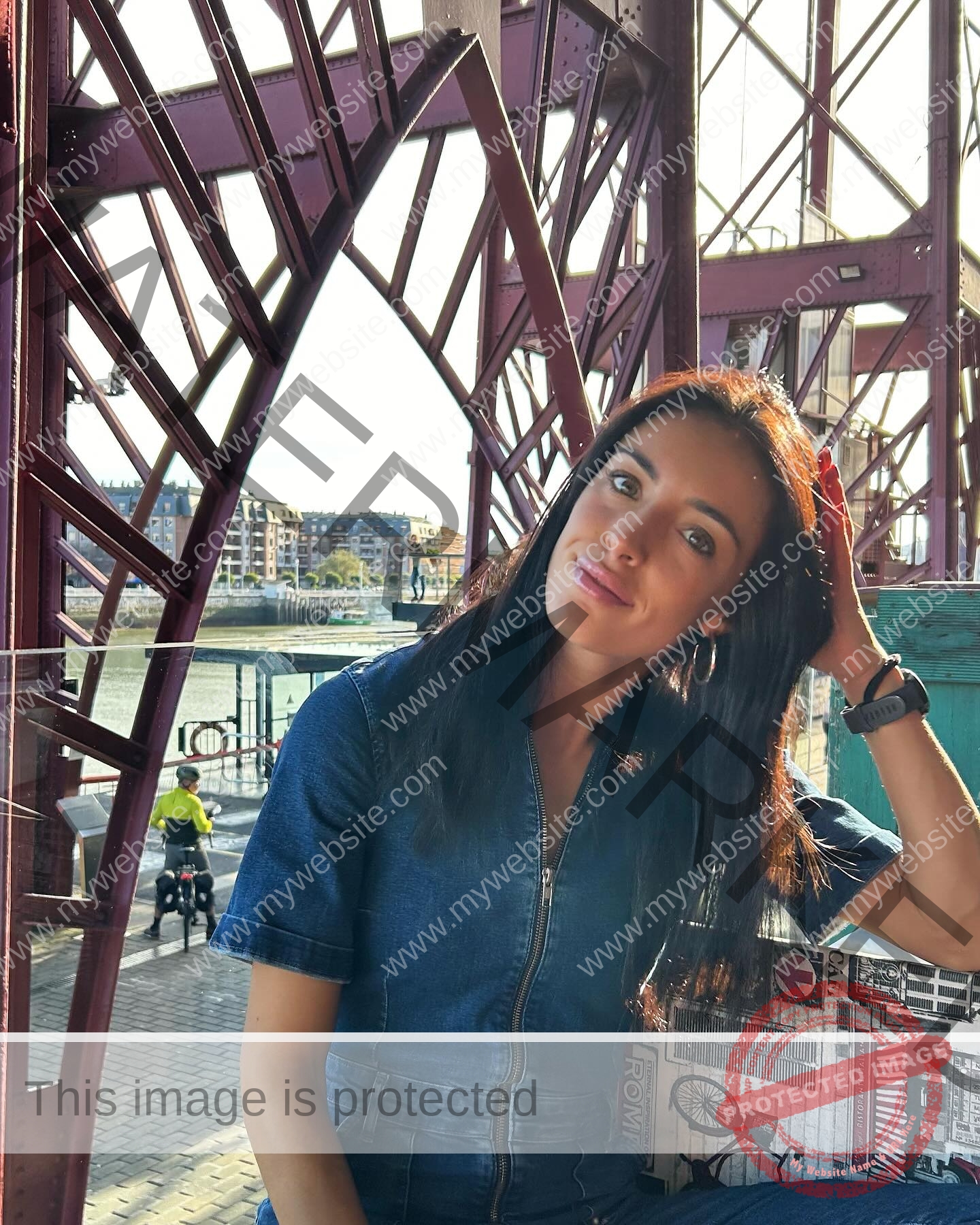ainhoa-idoiaga-spain-ainhoidoiaga-00354 Ainhoa Idoiaga, track athlete from Spain, sits in blue denim under a red bridge, touching her hair; city and cyclists in the background.