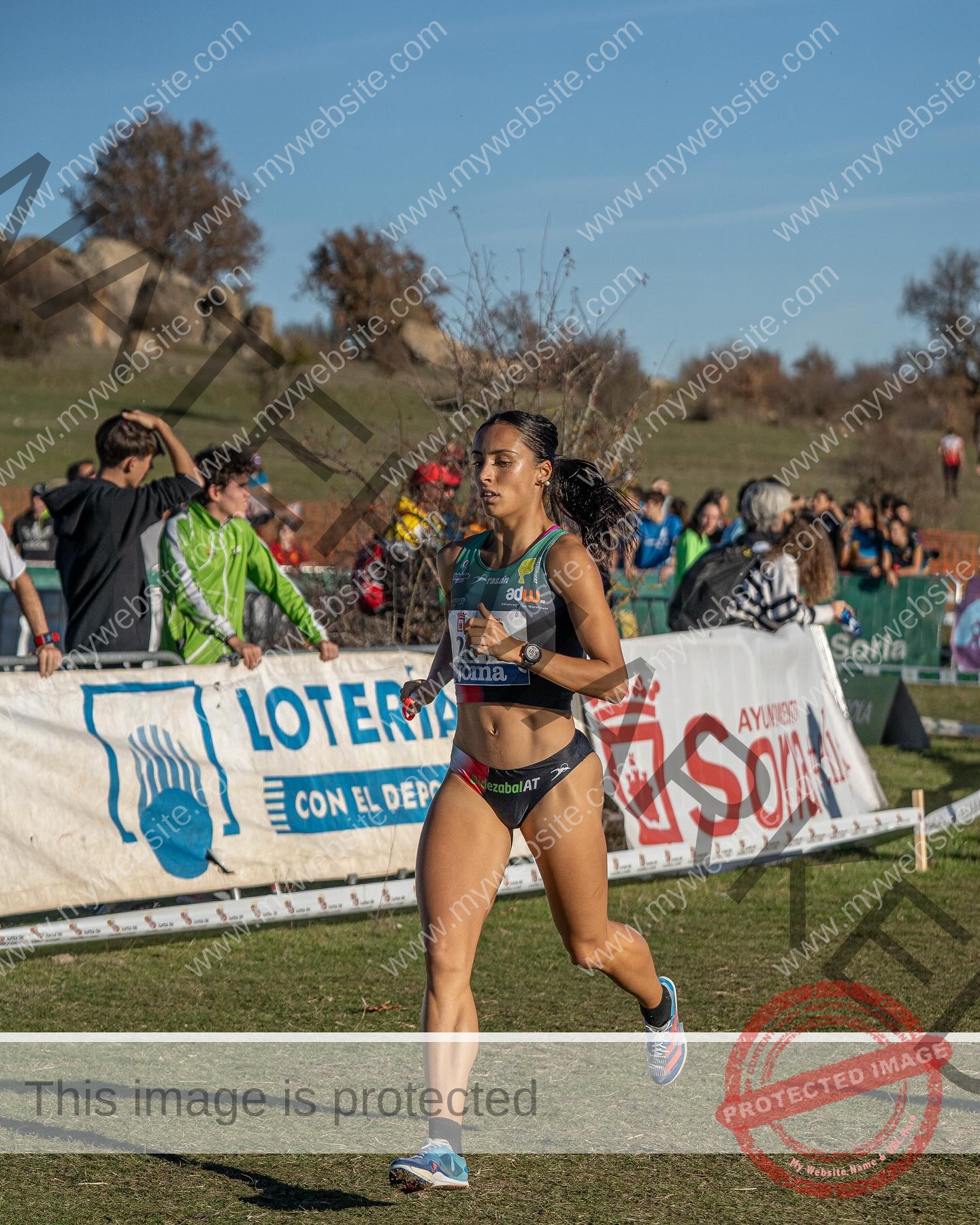 ainhoa-idoiaga-spain-ainhoidoiaga-00356 Ainhoa Idoiaga, track athlete from Spain, competes in athletic gear on grassy track, with spectators and banners under blue sky.