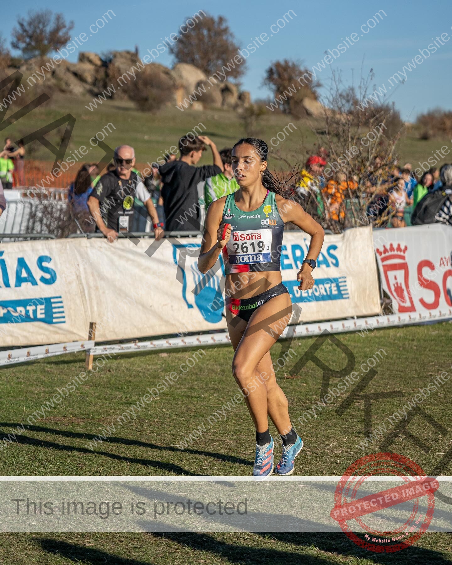 ainhoa-idoiaga-spain-ainhoidoiaga-00364 Ainhoa Idoiaga, track athlete from Spain, races on a grassy course in athletic gear, bib 2619, focused with spectators behind.