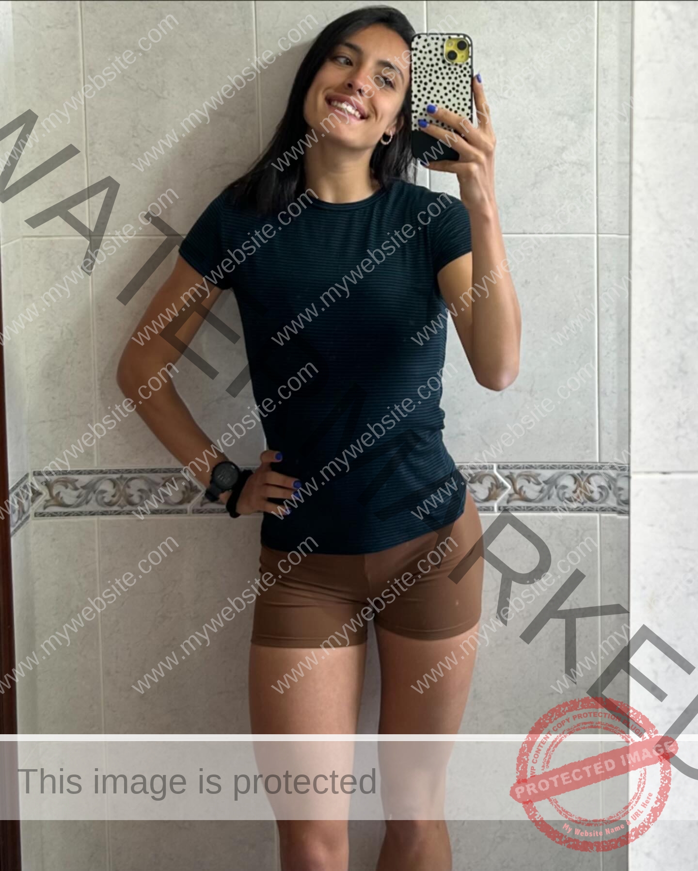ainhoa-idoiaga-spain-ainhoidoiaga-00375 Ainhoa Idoiaga, track athlete from Spain, with straight dark hair smiles taking a mirror selfie in a tiled bathroom, wearing black t-shirt and brown shorts.