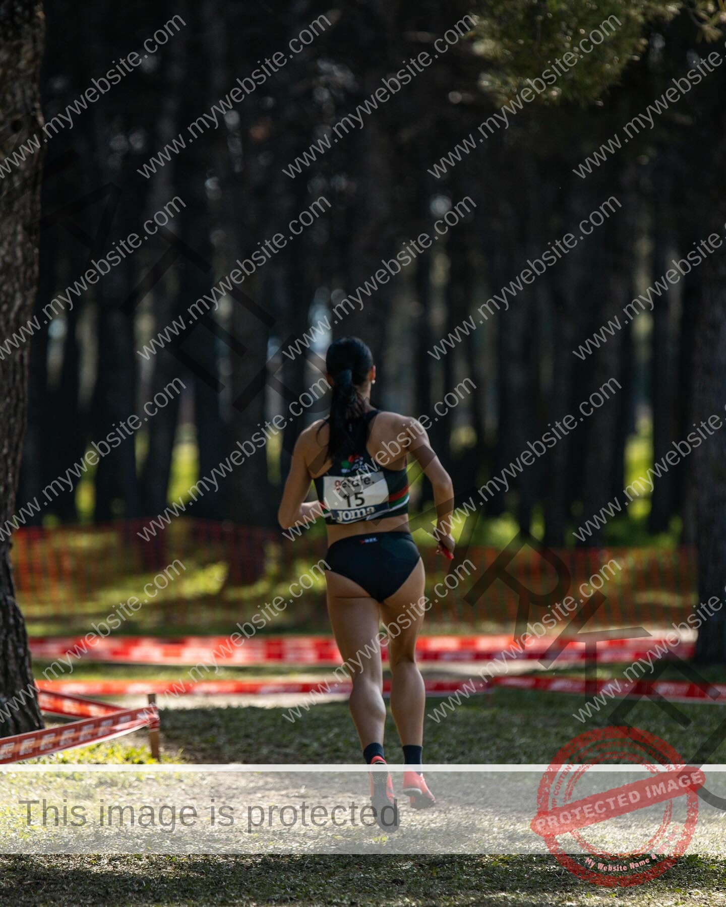 ainhoa-idoiaga-spain-ainhoidoiaga-00404 Ainhoa Idoiaga, track athlete from Spain, runs with a race bib along a forest path marked by red tape and sunlit trees.