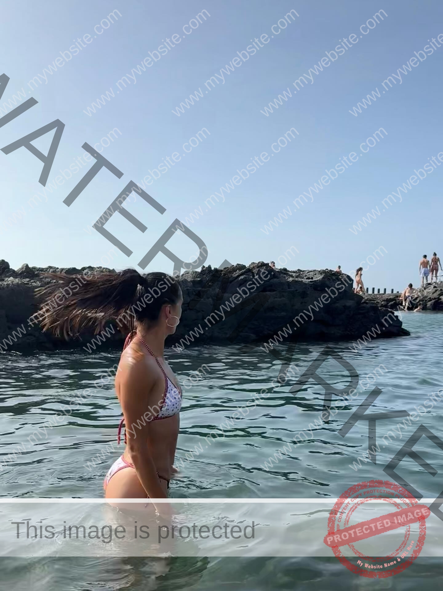 ainhoa-idoiaga-spain-ainhoidoiaga-00450 Ainhoa Idoiaga, track athlete from Spain, stands waist-deep in clear water wearing a pink and white bikini, looking toward a rocky shore.