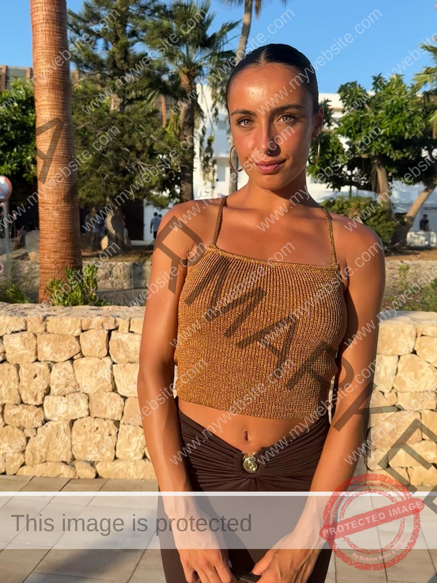 ainhoa-idoiaga-spain-ainhoidoiaga-00462 Ainhoa Idoiaga, track athlete from Spain, stands outdoors in gold crop top and brown skirt by a stone wall, looking relaxed.