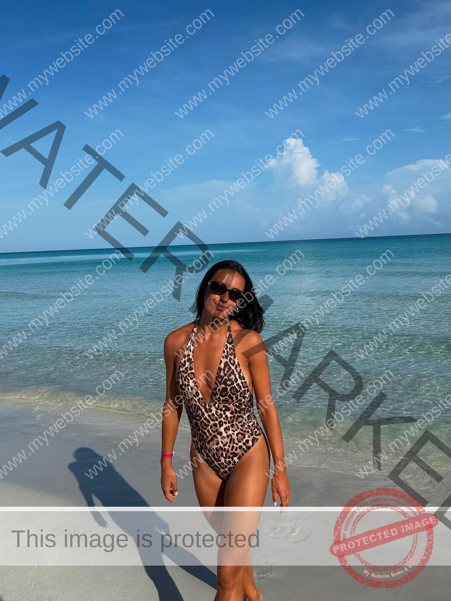 ainhoa-idoiaga-spain-ainhoidoiaga-00482 Ainhoa Idoiaga, track athlete from Spain, stands in a leopard print swimsuit and sunglasses on a sandy beach with clear blue water.