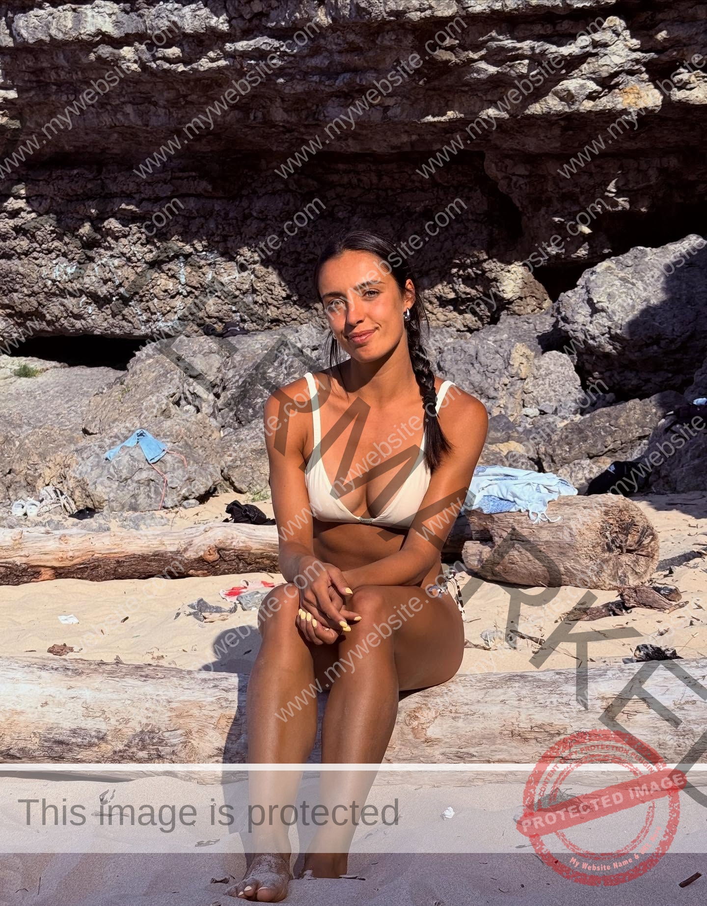 ainhoa-idoiaga-spain-ainhoidoiaga-00505 Ainhoa Idoiaga, track athlete from Spain, sits on a log in a light bikini on a sandy beach with rocky cliffs and sunlight.