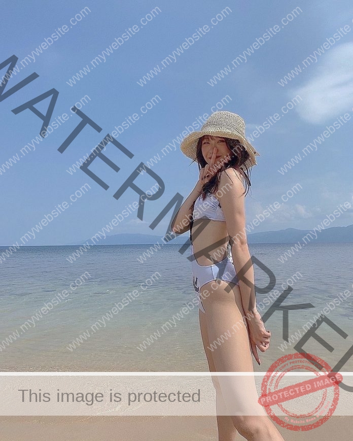 Akira Kobayashi, track athlete from Japan, stands in a white bikini and straw hat on a sunny beach by clear blue water, smiling.