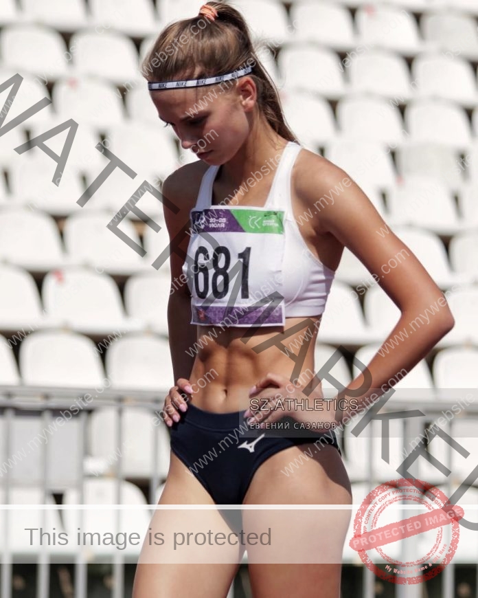 Alena Mikhaylovich, track athlete from Russia, stands head down on a stadium track in white sports bra and navy shorts, number 681.