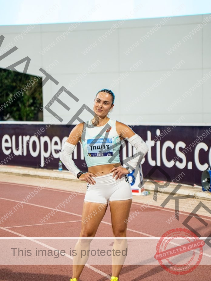 Alina Drutman Alina Drutman, a female Israeli track athlete, stands on the track hands on hips in white gear and race bib, European Athletics sign behind.