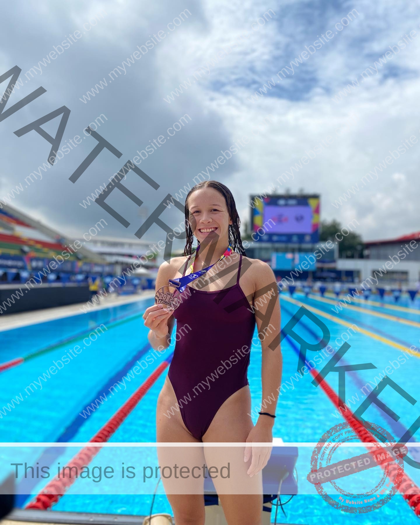 alondra-ortiz-costa-rica-alortizroman-437 Alondra Ortiz, wearing a maroon swimsuit, smiles and holds a medal by an outdoor pool; swim lanes and seating are in the background.