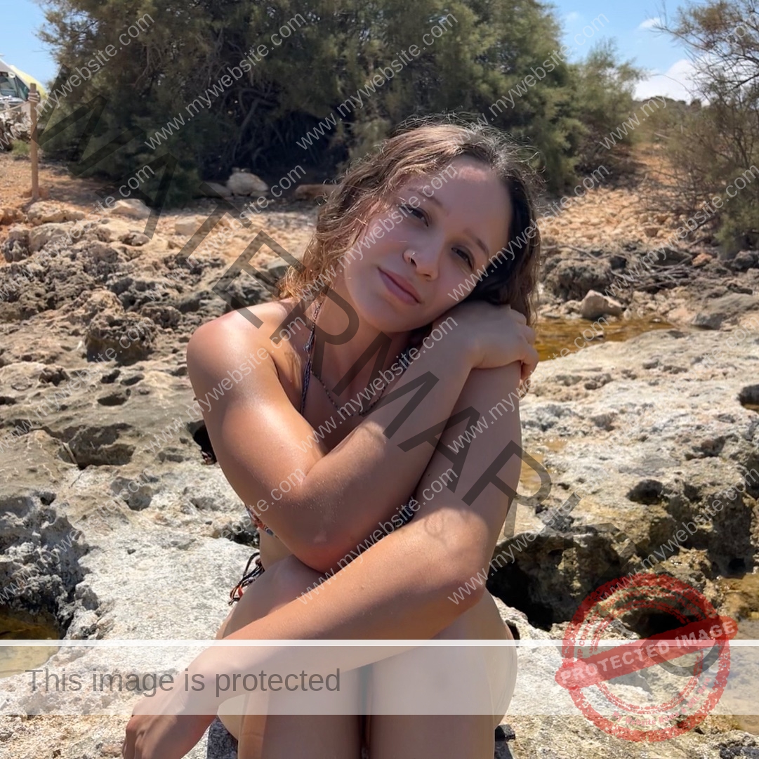alondra-ortiz-costa-rica-alortizroman-638 Alondra Ortiz sits on rocky ground in a bikini, knees hugged, smiling gently. Bushes and blue sky frame this serene Costa Rica scene.