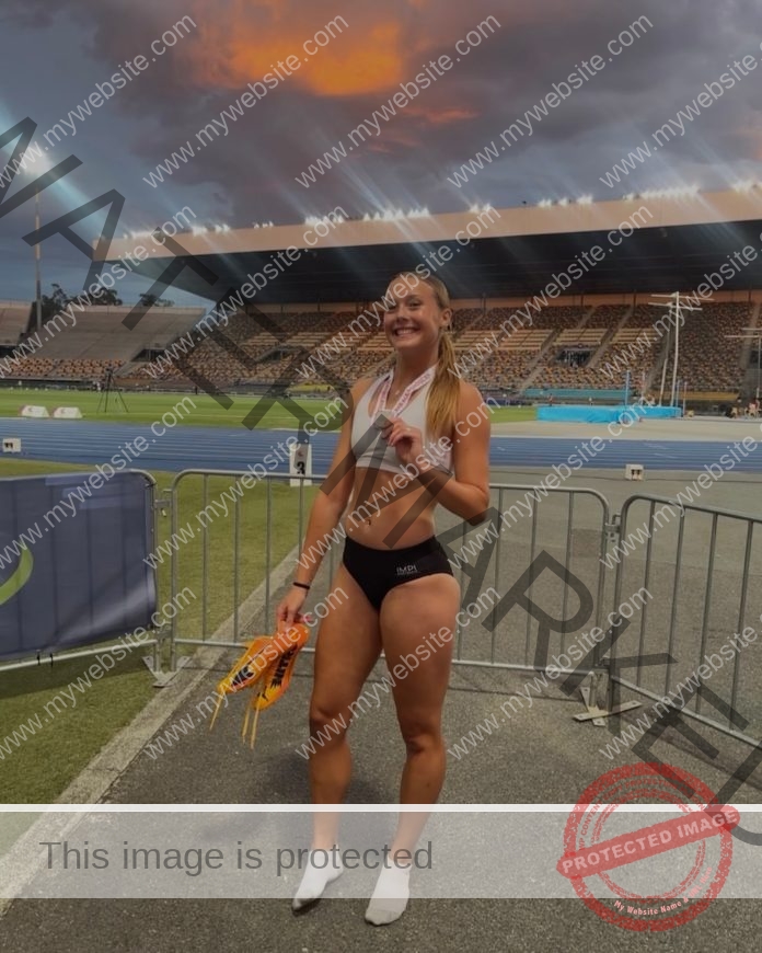 Alyssa McDonald A smiling track and field athlete, Alyssa McDonald of Australia, in white sports top and black shorts, holds medals and orange running spikes on a track near a stadium at sunset.