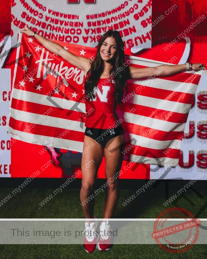 Amarae Krafka smiles in a red Nebraska Huskers track outfit, holding a team flag on green turf, with Huskers logos behind her.