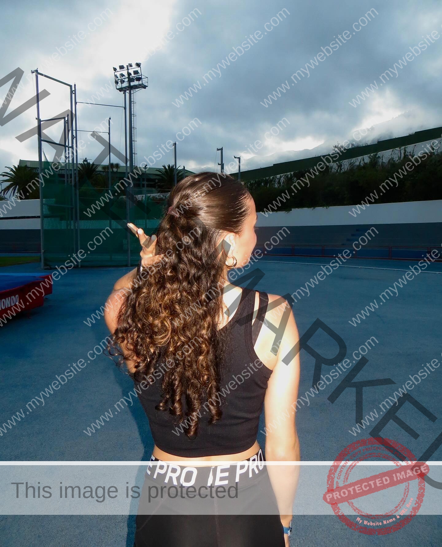 amira-never-germany-amira.never-02772 Amira Never, track star from Germany, with curly hair in a ponytail, in black sportswear on a blue track, raises her hand under clouds.