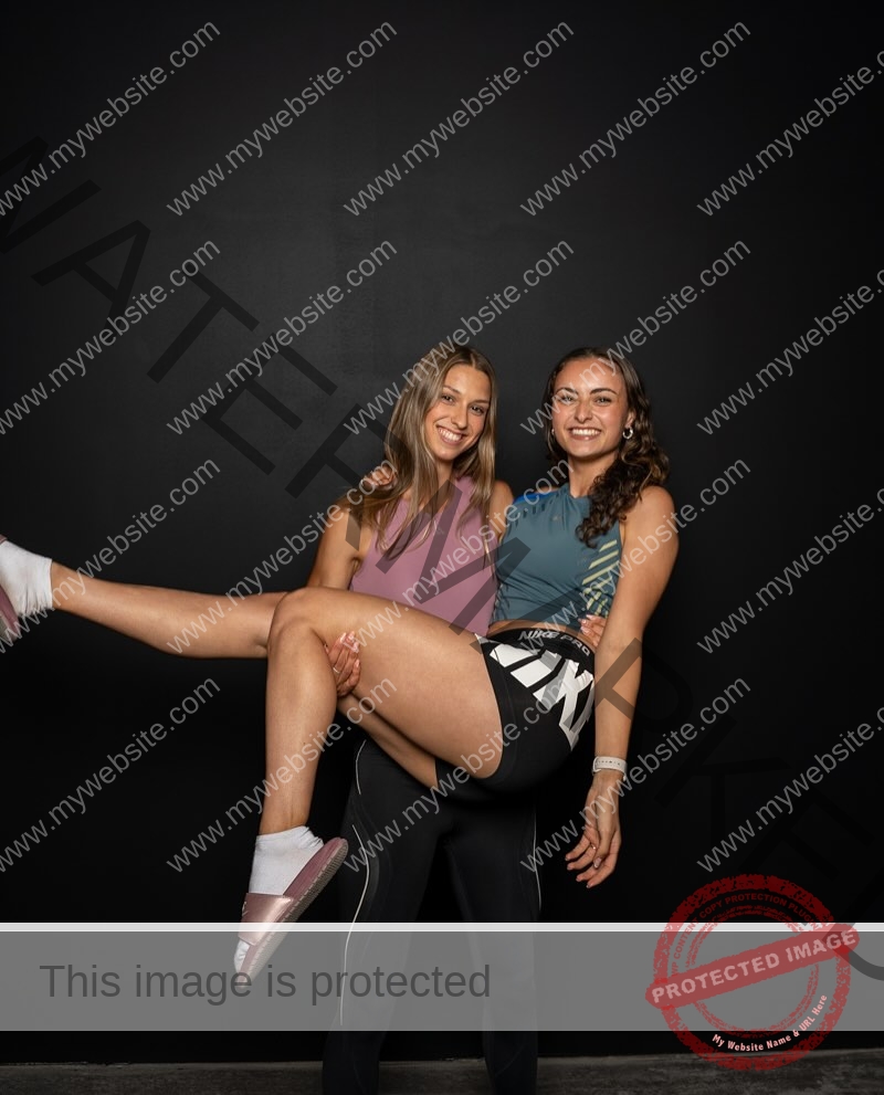 amira-never-germany-amira.never-02773 Amira Never, track star from Germany, two young women pose in front of a black background. One smiles carrying the other on her back.