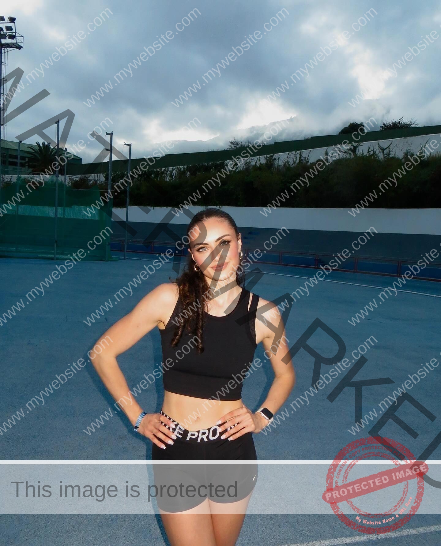 amira-never-germany-amira.never-02776 Amira Never, track star from Germany, stands on a cloudy outdoor track in black sportswear, smiling with hands on hips; trees and buildings behind.