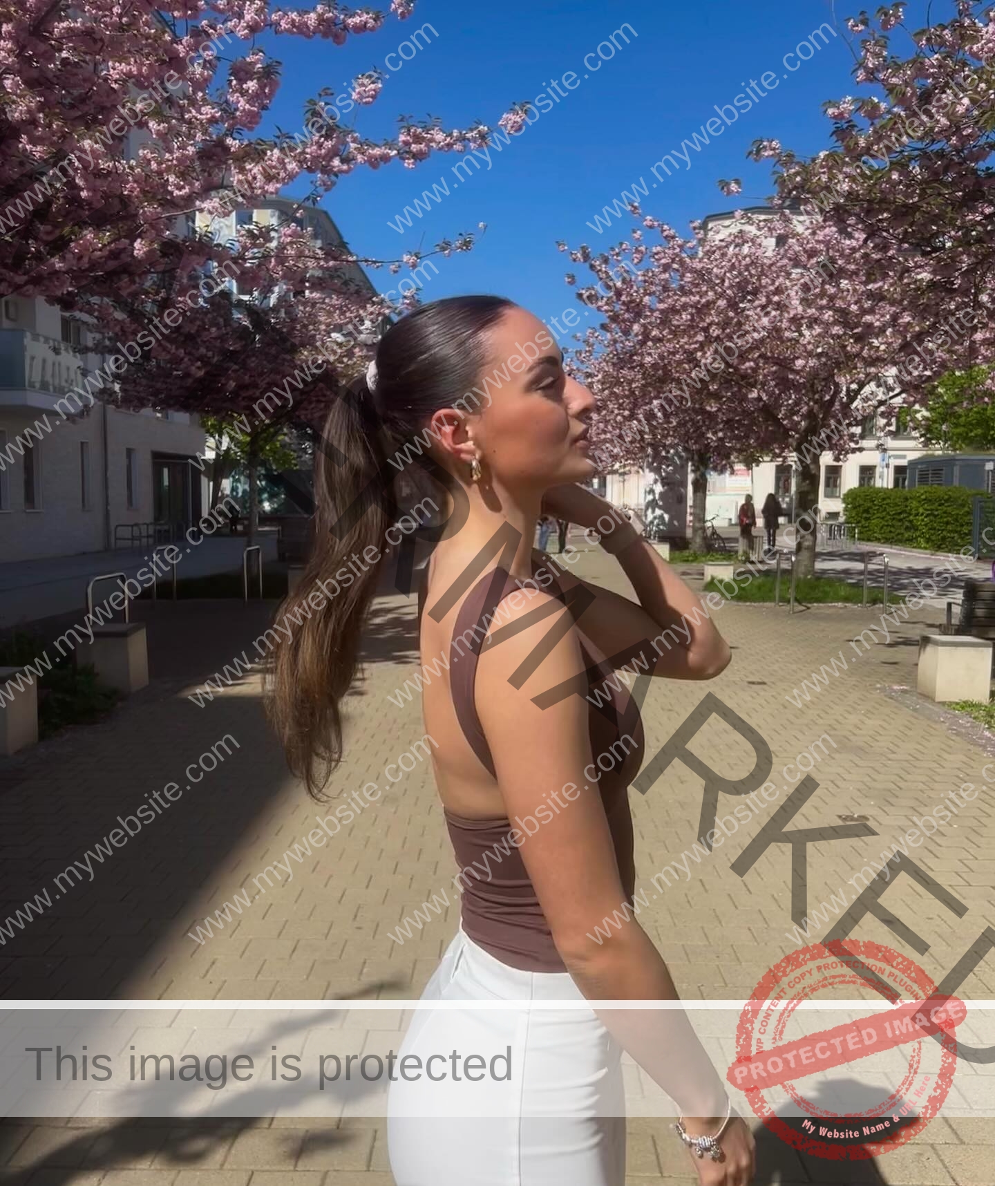 amira-never-germany-amira.never-02777 Amira Never, track star from Germany stands on a sunlit path lined with blooming cherry blossoms, in brown top and white pants.