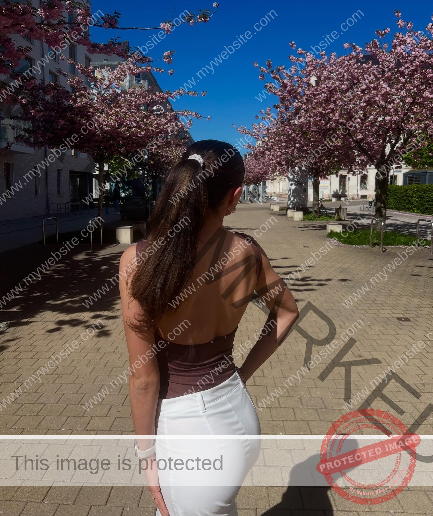 amira-never-germany-amira.never-02778 Amira Never, track star from Germany, with long brown hair in a ponytail and a backless top, walks past blooming cherry trees.