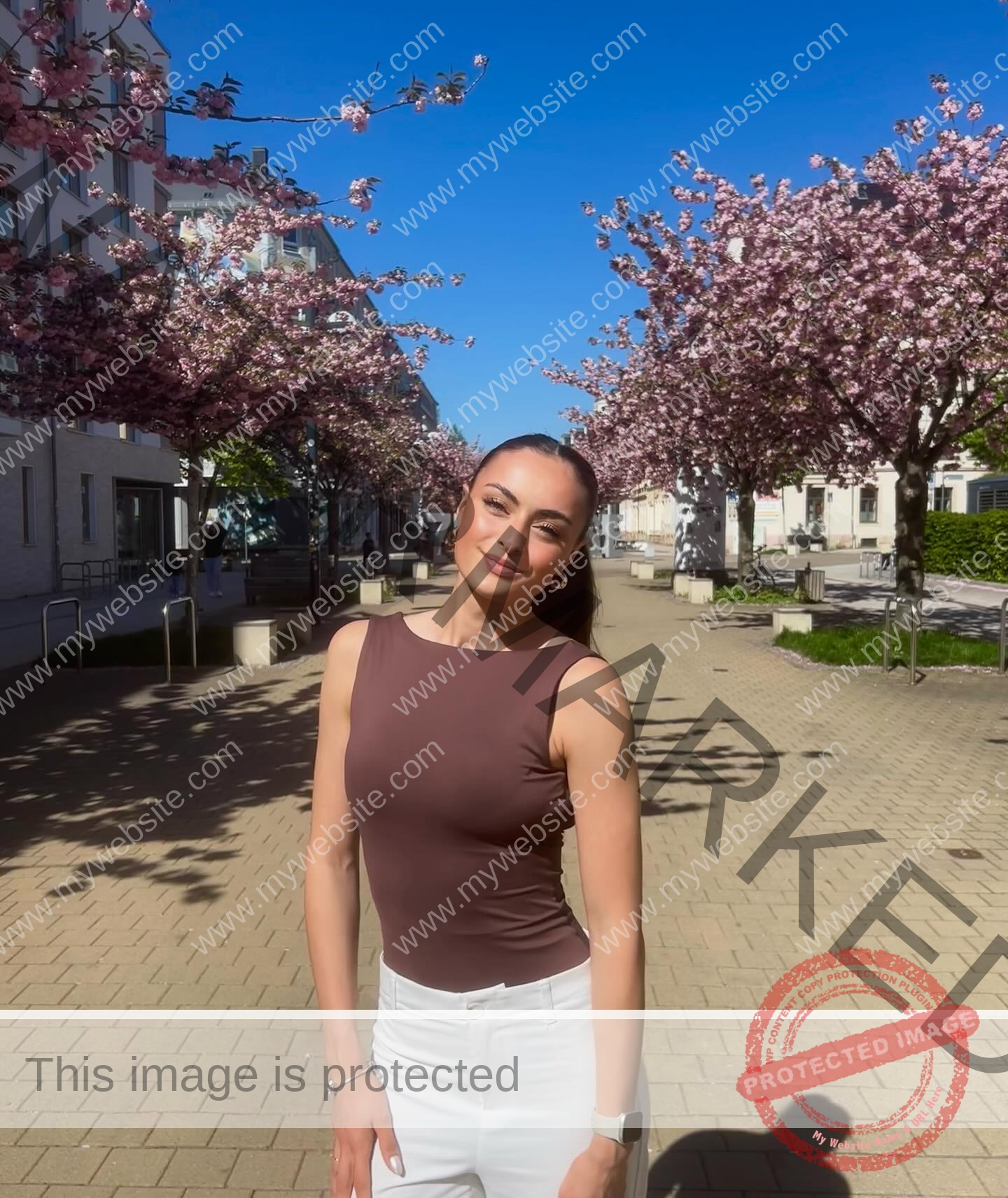 amira-never-germany-amira.never-02779 Amira Never, track star from Germany, in a brown sleeveless top and white pants, smiles on a sunny street with cherry blossoms.