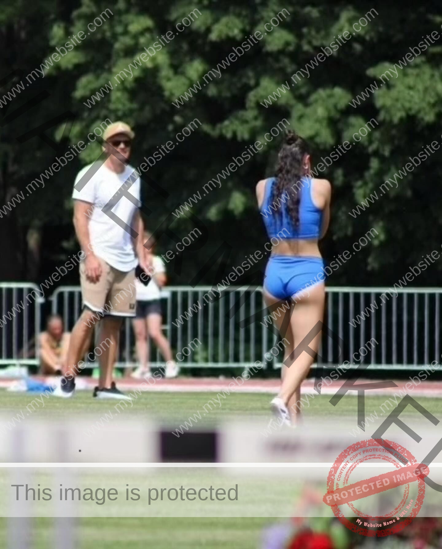amira-never-germany-amira.never-02782 Amira Never, track star from Germany, in blue sports gear walks on a track; a man stands nearby, with trees and a metal fence behind.