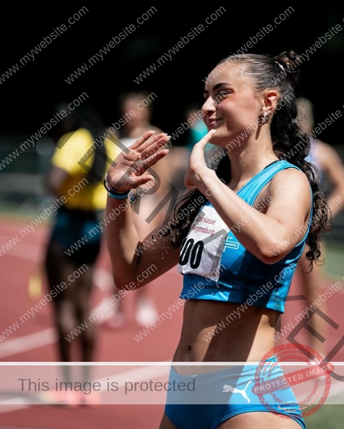 Amira Never, track star from Germany, in blue sportswear on a track, smiling and raising hands as blurred runners are in the background.