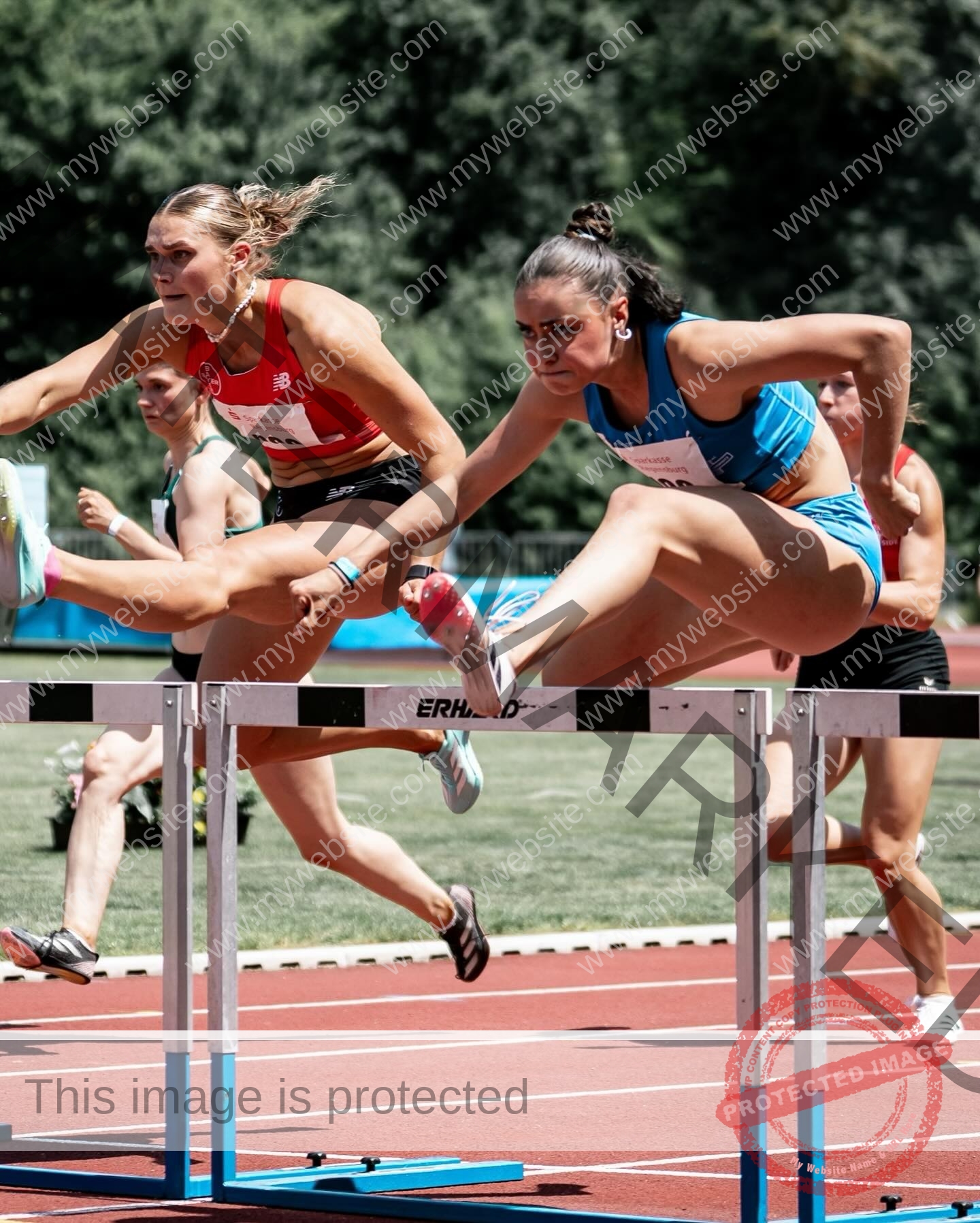 amira-never-germany-amira.never-02785 Amira Never, track star from Germany, and another athlete compete mid-jump in a hurdle race with others and greenery in the background.