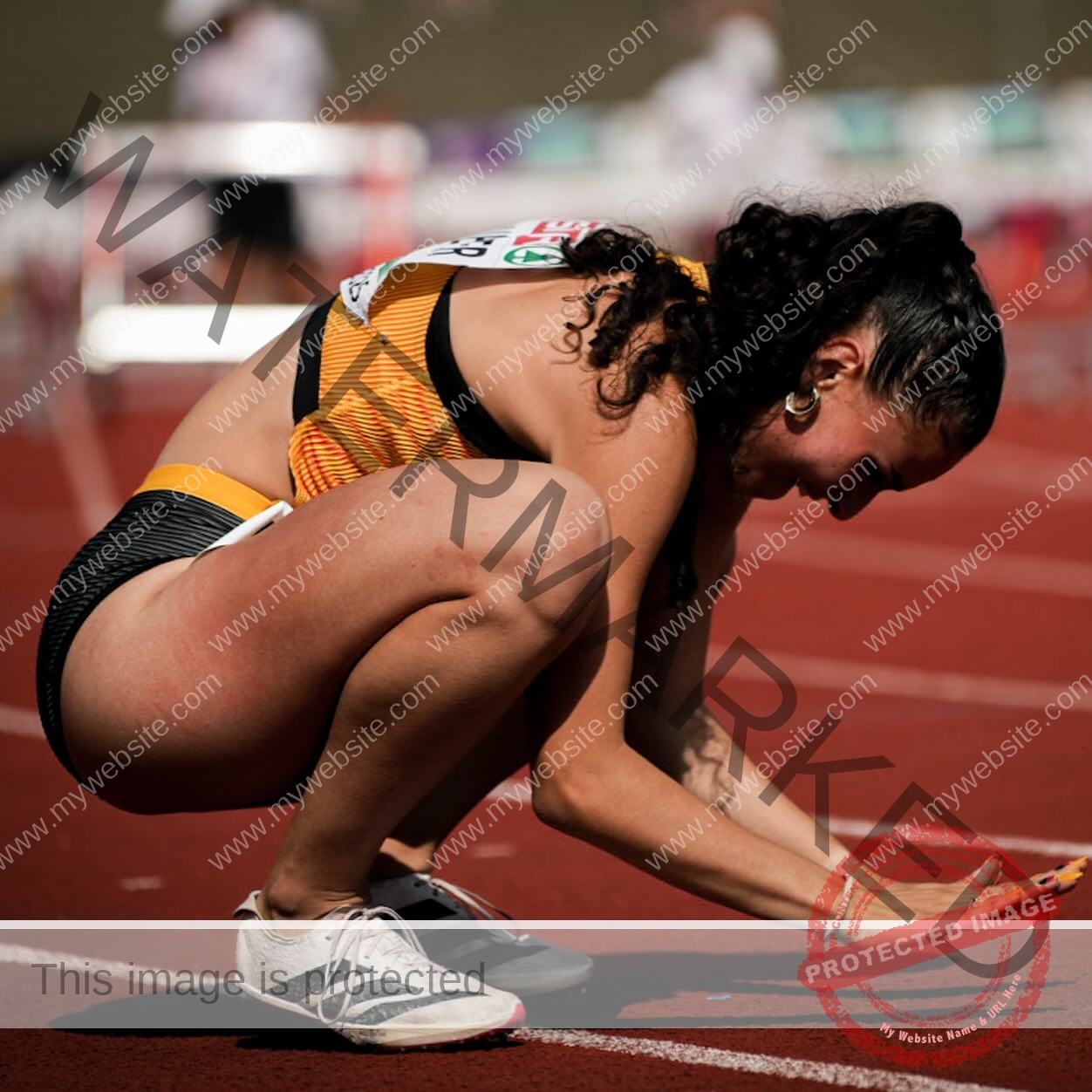 amira-never-germany-amira.never-02787 Amira Never, track star from Germany, in orange and black outfit crouches on a red track, smiling with one hand on the ground.