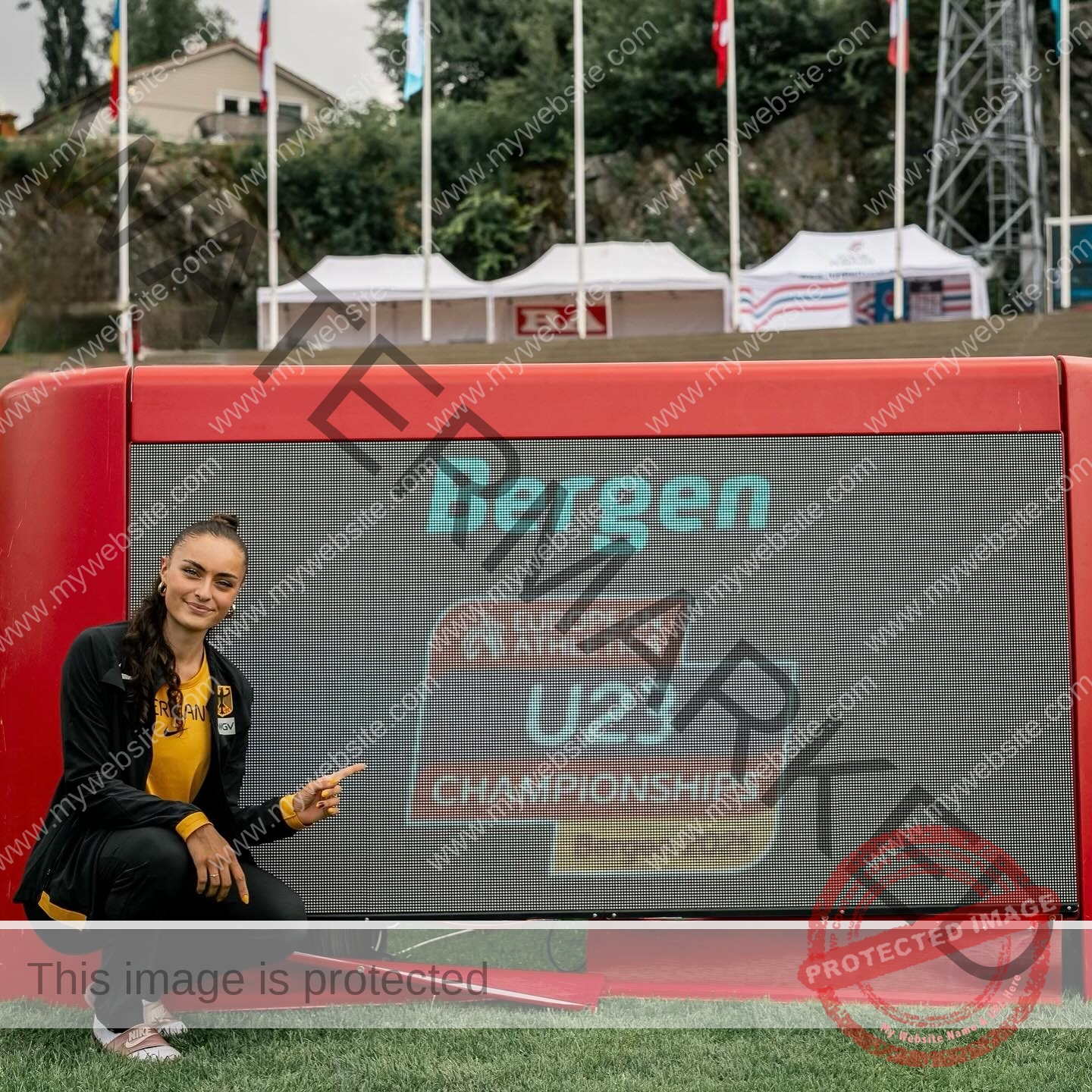 amira-never-germany-amira.never-02790 Amira Never, track star from Germany crouches on grass and points at a large digital screen at an outdoor sports venue.