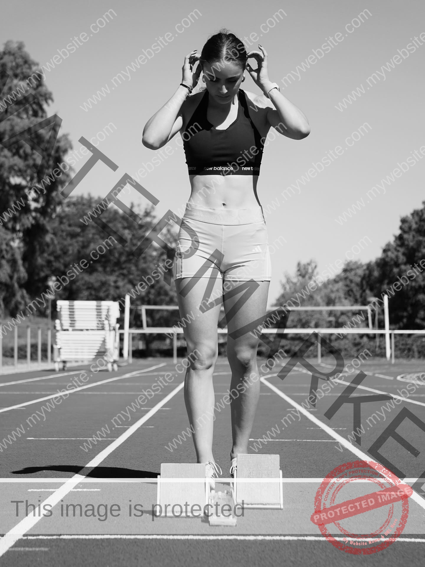 amira-never-germany-amira.never-02793 Amira Never, track star from Germany, stands on a running track in athletic wear, adjusting her hair over starting blocks; black and white.