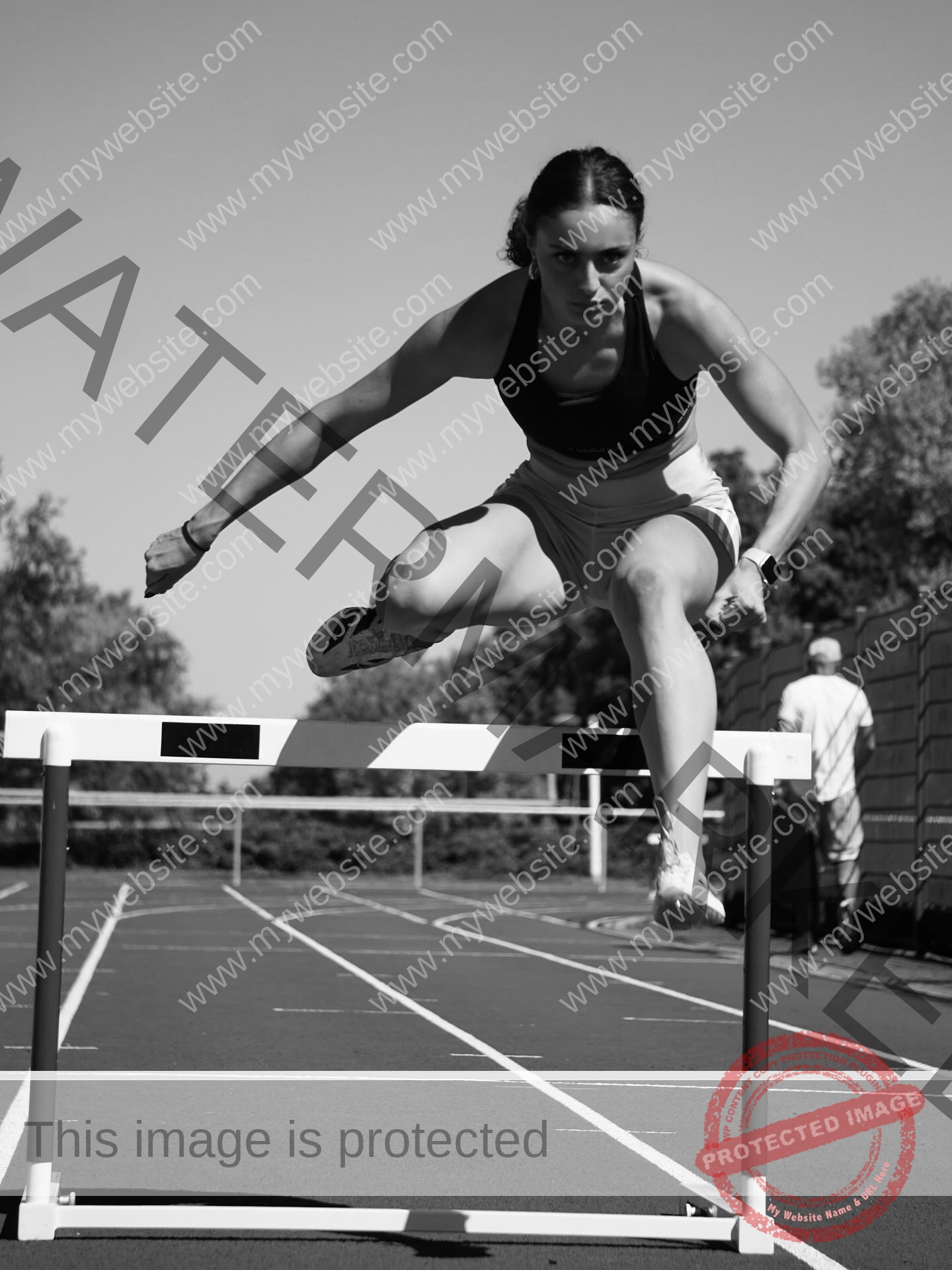 amira-never-germany-amira.never-02795 Amira Never, track star from Germany leaps over a hurdle mid-jump on an outdoor track; black and white, with blurred figure behind.