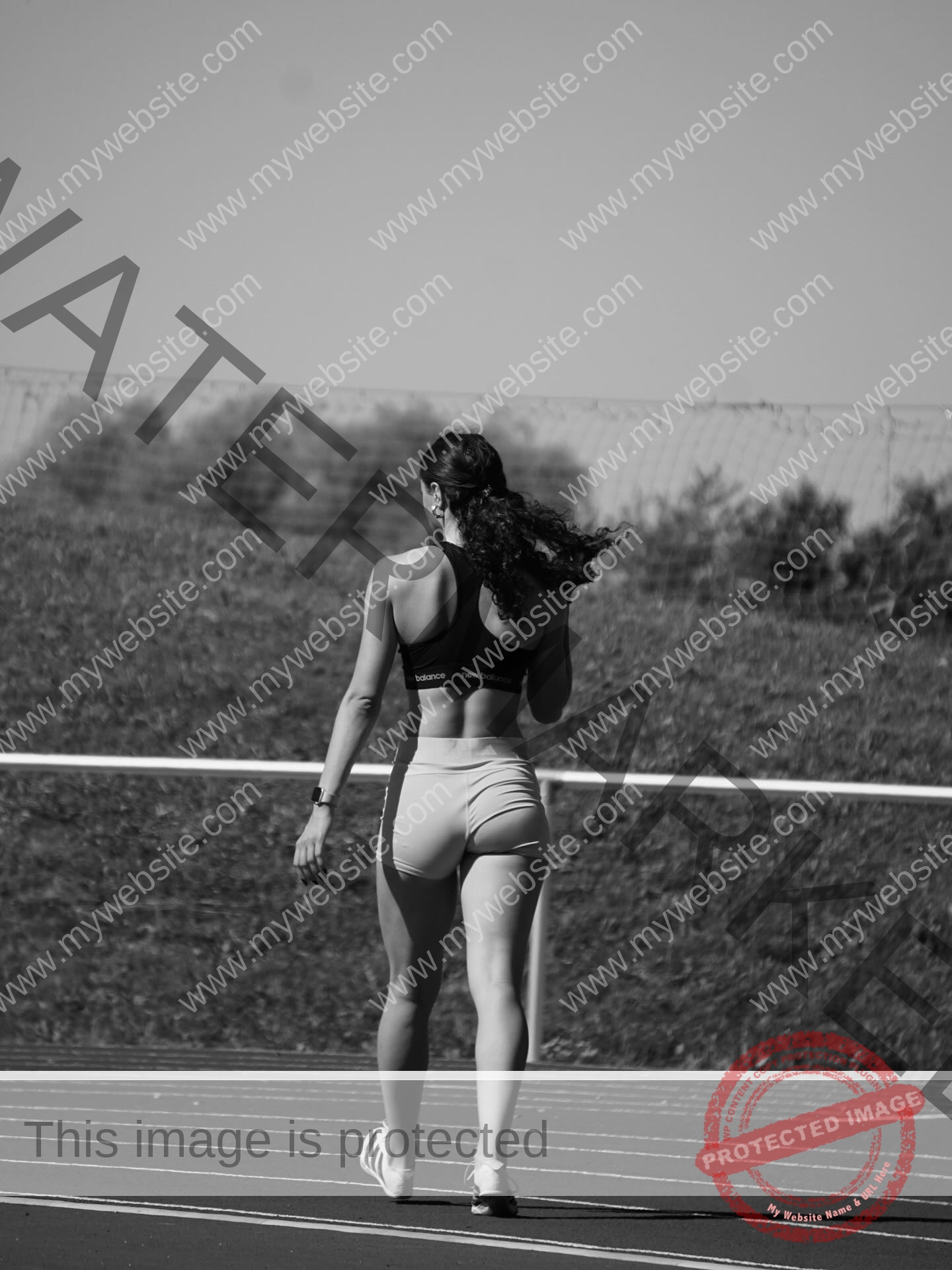 amira-never-germany-amira.never-02798 Amira Never, track star from Germany walks alone on a black and white outdoor track, hair tied, grassy terrain and fence behind.